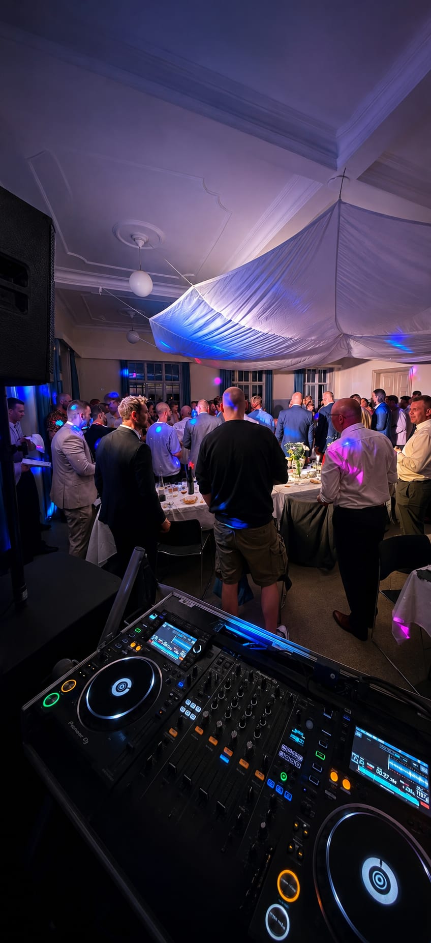 DJ mixing deck overlooking guests mingling at a small indoor event with draped ceiling decor.