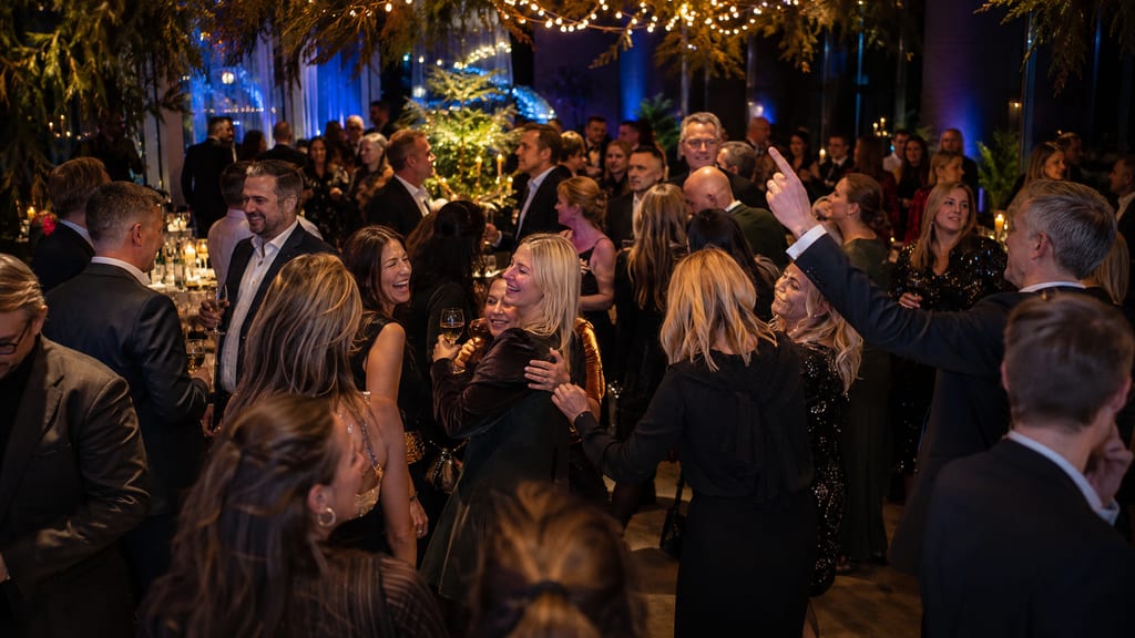 People in formal attire mingle and laugh at a festive Christmas party with fairy lights and a tree.